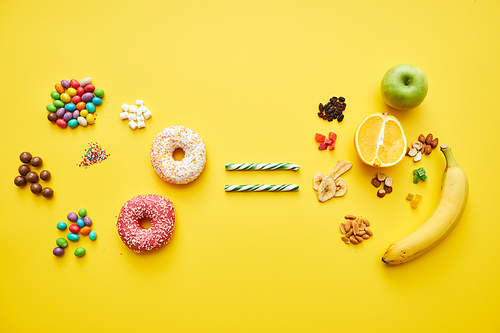 High angle view of unhealthy candies and sweet doughnuts being equated with fresh and dried fruits on yellow background
