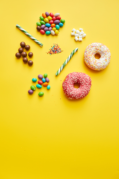 Directly above view of various sweets and candies such as jellybeans, sprinkles, marshmallows, candy canes and doughnuts on yellow background