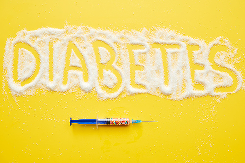 High angle view of plastic syringe with sweet sprinkles and diabetes word written on granulated sugar, yellow background