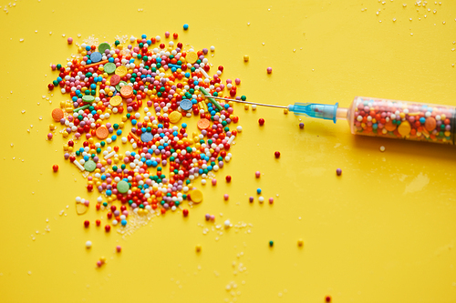 Close-up of splattering sweet sprinkles and needle of medical syringe on bright yellow background