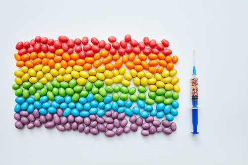 Drugs and equality flag: plastic syringe with sweet sprinkles fitted with needle and multi-colored flag made of jellybeans, knolling composition