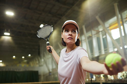Determined attractive young female tennis player in cap standing on court and swinging arm with racket while making serving