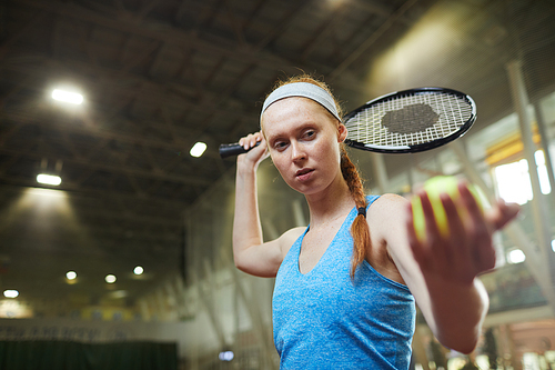 Serious focused female professional tennis player in blue shirt and headband standing on indoor court and serving ball while starting match