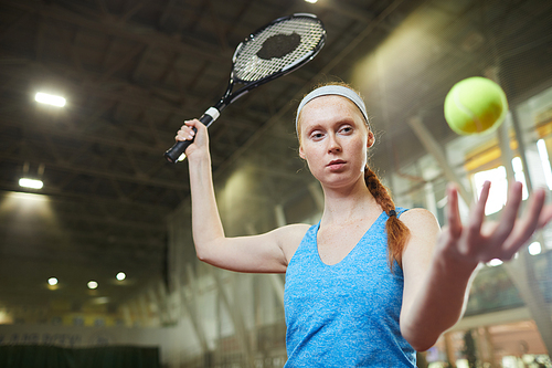 Serious confident redhead female tennis player with braid standing on indoor court and throwing ball while practicing hit