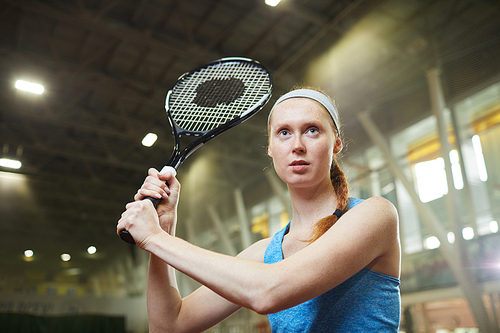 Concentrated young woman with red braid standing on spacious court and gripping tennis racket to hit ball