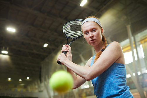 Portrait of serious redhead female tennis player in sports headband focused on movement of ball hitting tennis ball on court