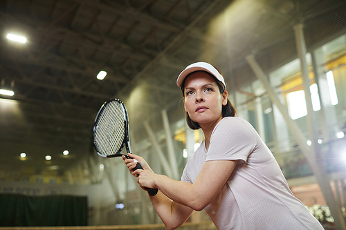Serious skilled young woman in cap standing on indoor court and holding racquet while catching tennis ball with racket