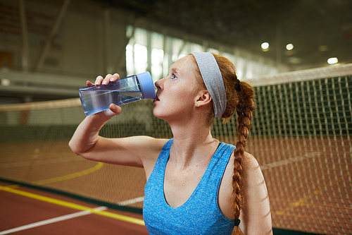 Sporty young woman with red braid standing by tennis net and drinking refreshing water from bottle after training