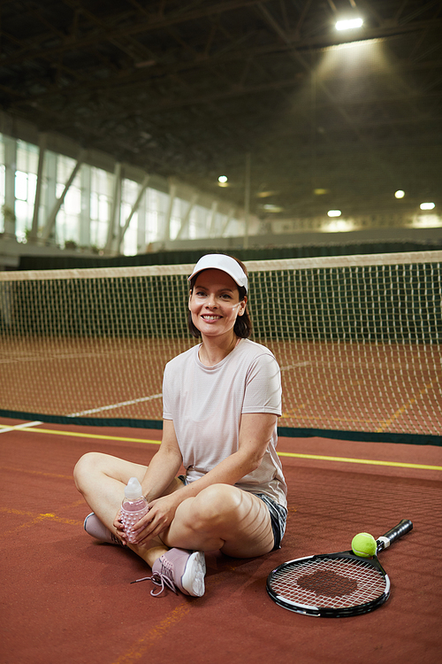 Portrait of happy sporty young woman in cap sitting with crossed legs on tennis court floor with personal racket and holding water bottle