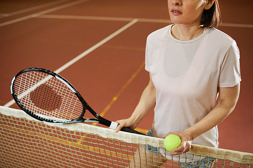 Close-up of content woman in comfortable tshirt standing by net and holding ball and racket while discussing tennis rules with competitor