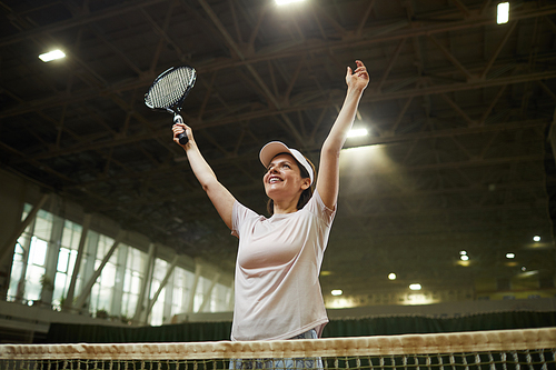 Cheerful excited young woman in cap standing by net and raising arms up while celebrating winning in tennis match