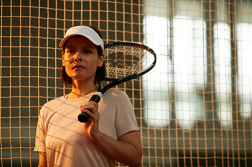 Portrait of serious confident female tennis player in cap standing against net on court and keeping tennis racquet on shoulder