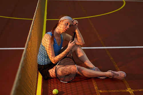 Thoughtful young redhead woman with braid exhausted after tennis match sitting on floor at dark tennis court and holding racket