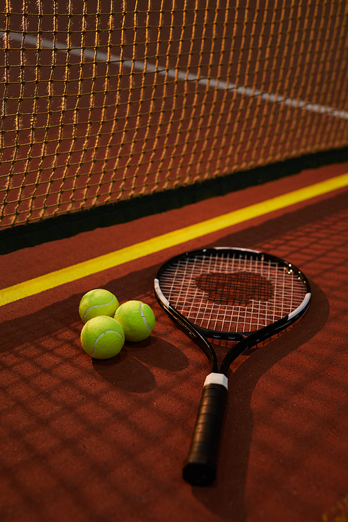Close-up of well-string tension racket and three yellow balls lying on dark tennis court, shadow of net effect