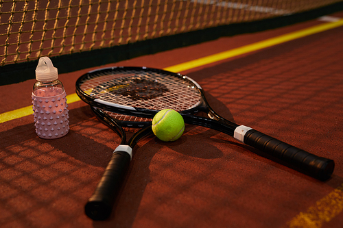 Close-up of modern tennis rackets, ball and pink bottle of water lying by net on floor in dark court, copy space