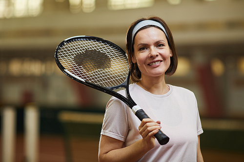 Portrait of happy energetic middle-aged woman in sports headband standing on court and holding tennis racket on shoulder