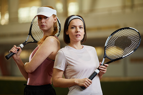 Sports team of female tennis players with rackets standing back to back on indoor court and looking at camera