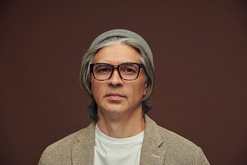 Portrait of cheerful handsome mature man in eyeglasses and gray hat against brown background, positive emotion concept