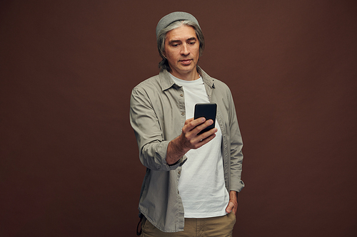 Positive hipster mature man in beanie hat standing against brown background and chatting by phone