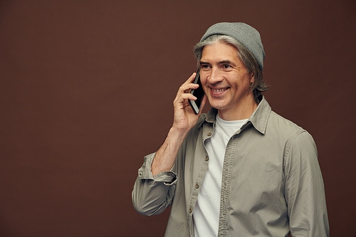 Portrait of smiling hipster aged man in gray hat standing against isolated brown background