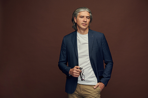 Portrait of serious gray-haired business executive in smart casual jacket holding eyeglasses against isolated background