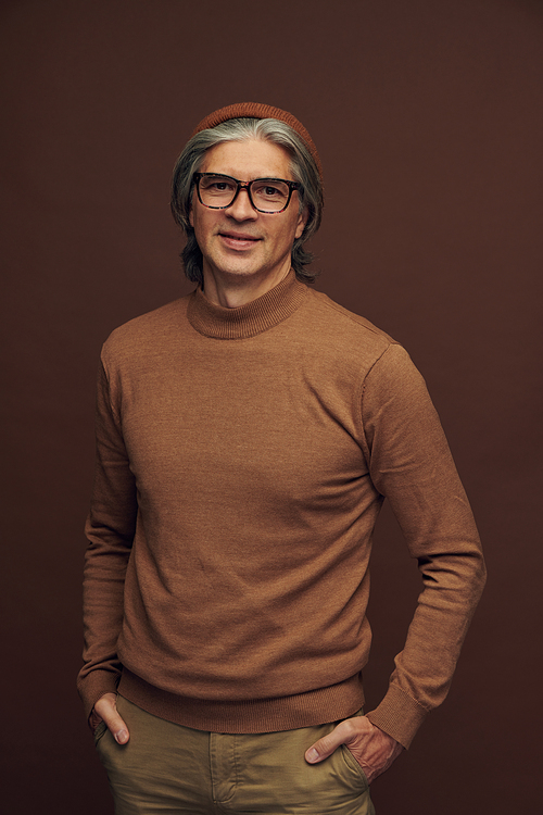 Portrait of content creative mature man in beanie hat and glasses posing against brown background