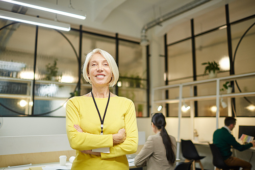 Cheerful confident attractive mature lady in yellow sweater standing in modern office and looking at camera