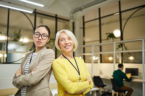 Positive successful multi-ethnic ladies with badges on necks standing back to back in modern office and looking at camera