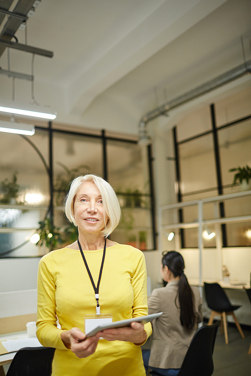 Smiling blond-haired mature woman wearing yellow sweater holding clipboard and looking at camera while working in modern company