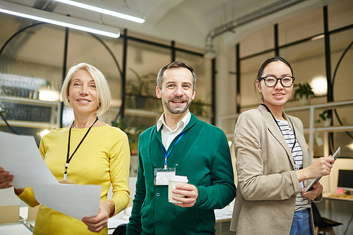 Group of cheerful multi-ethnic business team with badges standing in modern office and looking at camera