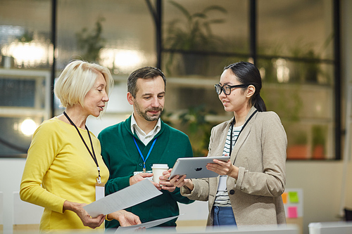 Smiling attractive young Asian woman in glasses showing tablet to colleagues and offering strategy