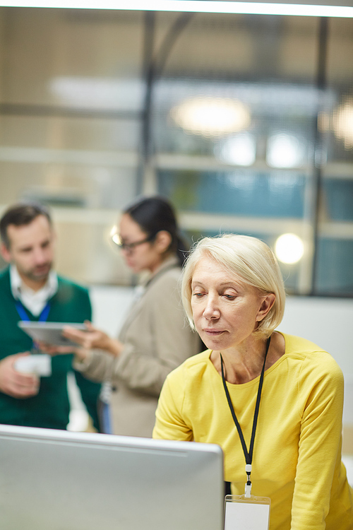 Serious thoughtful mature lady with badge on neck standing in front of computer and checking information