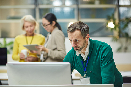 Serious frowning middle-aged man in green cardigan standing at table and analyzing project plan on computer