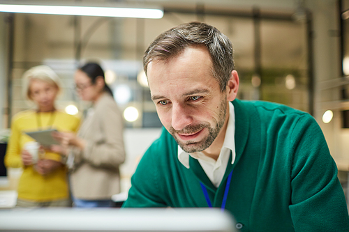 Smiling satisfied handsome businessman in green cardigan standing in office and viewing online statistics on computer