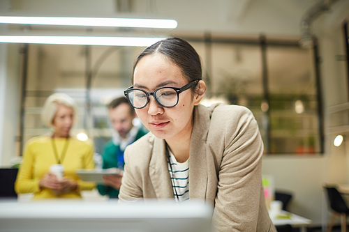 Smiling smart Asian woman in glasses standing in front of computer and working with data in office