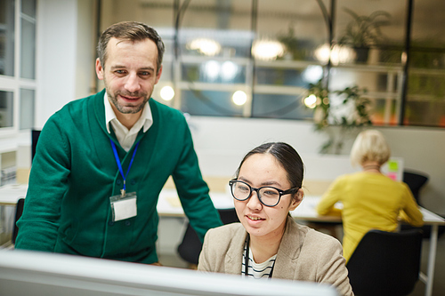 Confident handsome business mentor with badge standing at colleague and examining work of intern in office