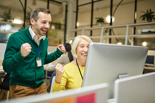 Excited successful colleagues in bright sweaters making yes gesture while looking at computer screen, they celebrating success