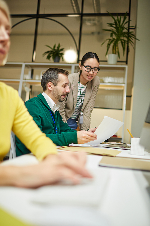 Smiling young Asian woman in glasses listening to colleagues advice while they examining papers in office