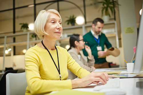 Content confident mature lady with blond hair sitting at desk and typing on computer keyboard in office