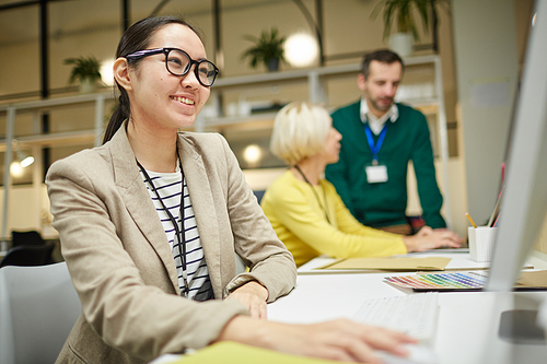 Smiling attractive young Asian manager in glasses sitting at table and using computer while analyzing report