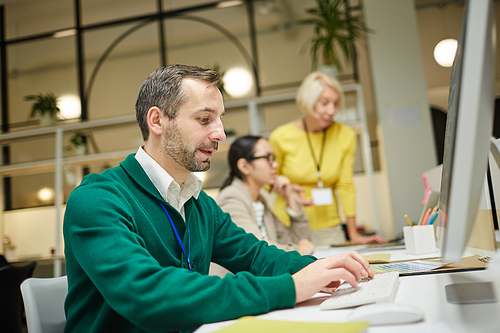 Serious modern programmer in green cardigan sitting at table and coding information on computer in office