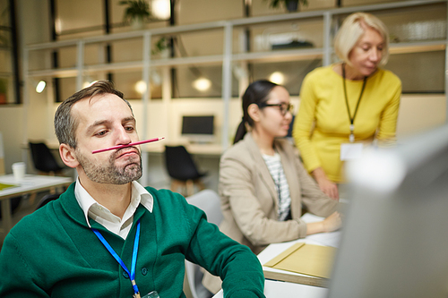 Careless handsome middle-aged employee with stubble keeping pencil on lip while wasting time in office