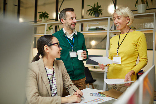 Group of creative colleagues in casual clothing discussing design and statistics while working with papers in modern office