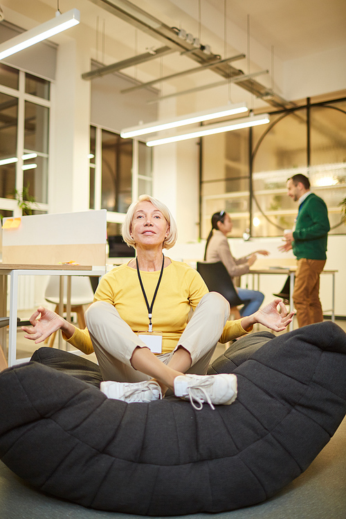 Content attractive blond-haired lady in yellow sweater wearing badge on neck sitting with crossed legs and practicing yoga in office