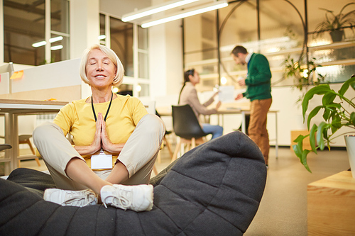 Smiling satisfied blond-haired woman with badge sitting with crossed legs on gray bean bag and relaxing with yoga at workplace