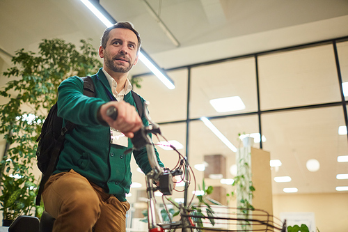 Modern middle-aged entrepreneur with backpack leaving office on electric bicycle