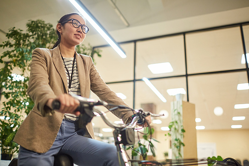 Pretty smiling Asian businessman in glasses enjoying riding on modern electric bicycle in office