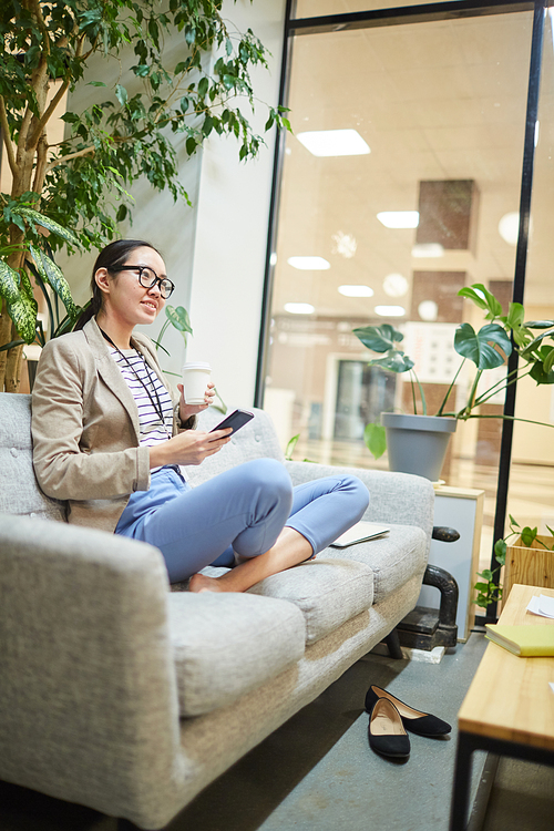 Pretty smiling Asian businesswoman sitting on comfortable sofa in staff room in office, drinking coffee and checking phone