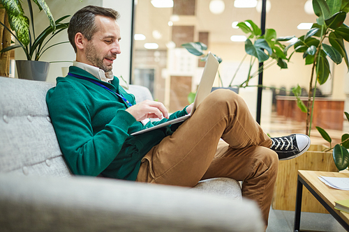 Smiling mature businessman sitting on comfy couch in his office and answering e-mails of clients on laptop