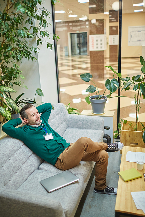 Smiling mature businessman leaning back on sofa in staff room and enjoying break after successful work